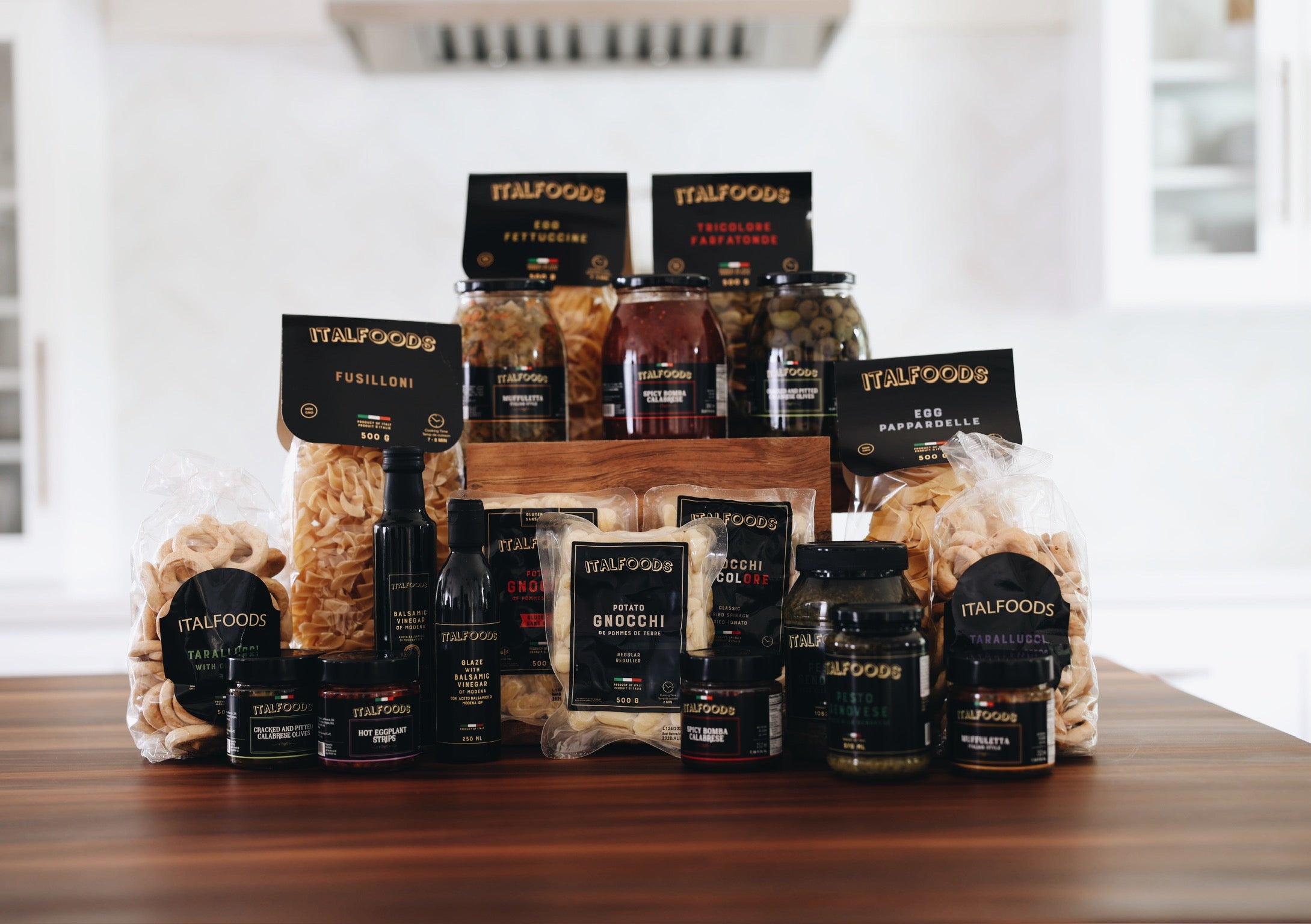 Assorted food products from ITALFOODS on a wooden table with a blurred kitchen background.