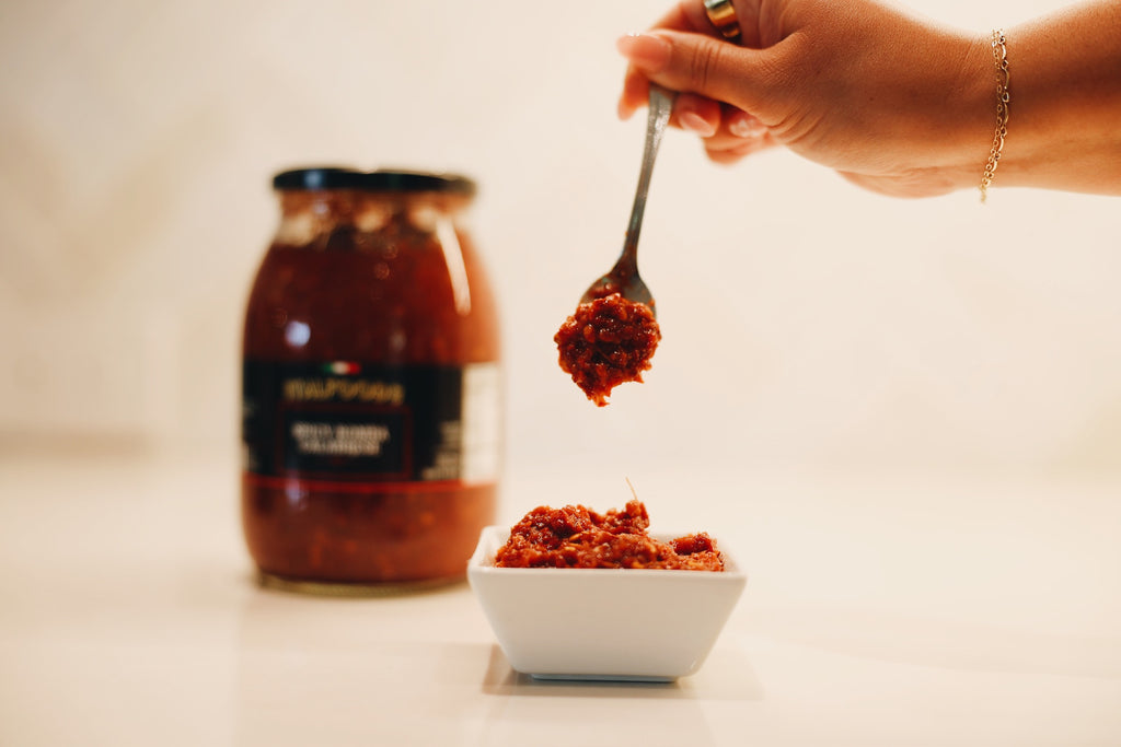 Hand holding a spoon with red paste over a small white bowl, jar of red paste in the background.