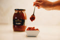 Hand holding a spoon with red paste over a small white bowl, jar of red paste in the background.